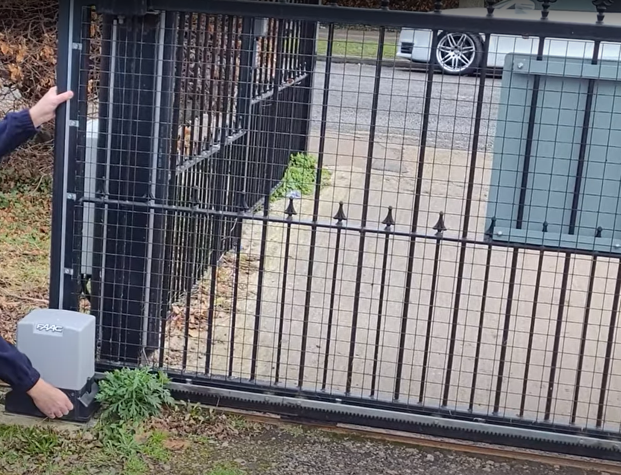 A residential gate repair technician is examining a partially open gate mechanism in front of a house driveway.